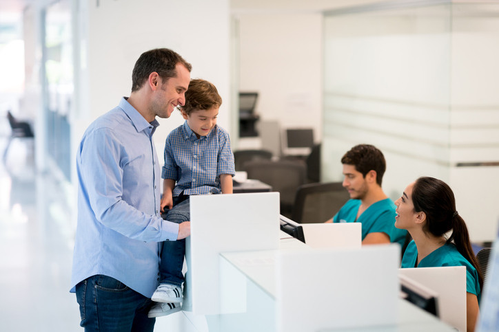 Father and son at general practice reception desk, talking to nurse.